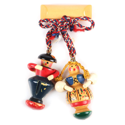 Vintage Bakelite Martha Sleeper bar pin with dangling painted carved German figures. Celluloid arms, waxed string braids, and braided floss bow. Man is missing a plume piece from his cap. 3 1/2" H x 2 1/2" W
