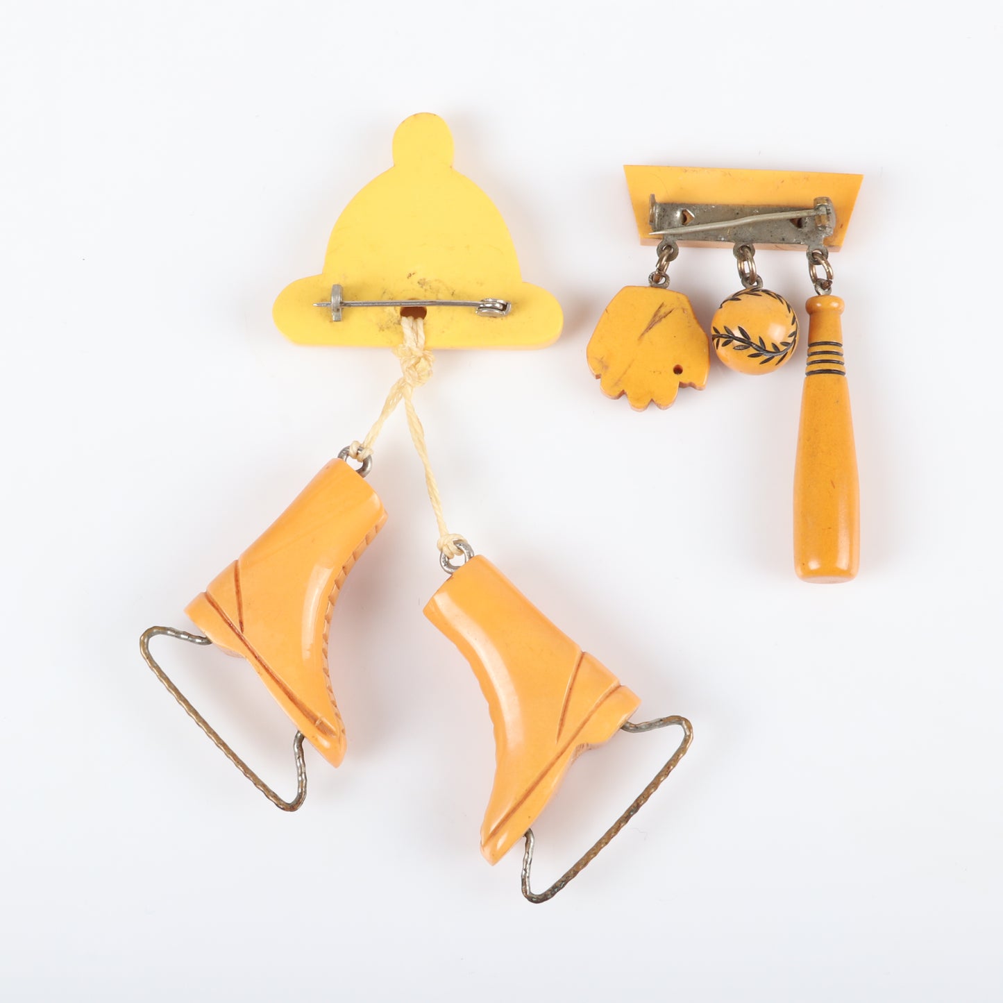 Vintage Bakelite 2pc. sports theme dangle charm pins. Carved butterscotch ice skates and cap, and baseball, bat and glove charms on bar pin. 4 1/4" H x 2 1/2" W (one)